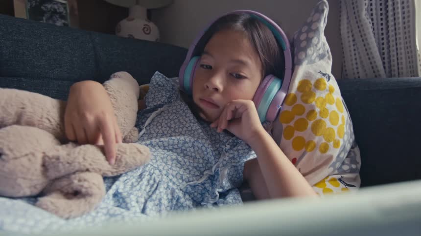 A young girl wearing headphones relaxes on a couch with her teddy bear while watching a tablet