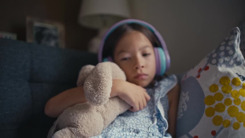 A young girl with headphones quietly watches a tablet while cuddling her stuffed animal on the couch