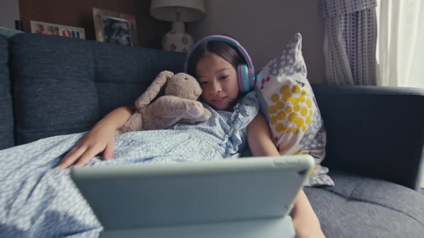 A young girl wearing headphones relaxes on a couch with her stuffed toy, attentively watching a video on a tablet computer