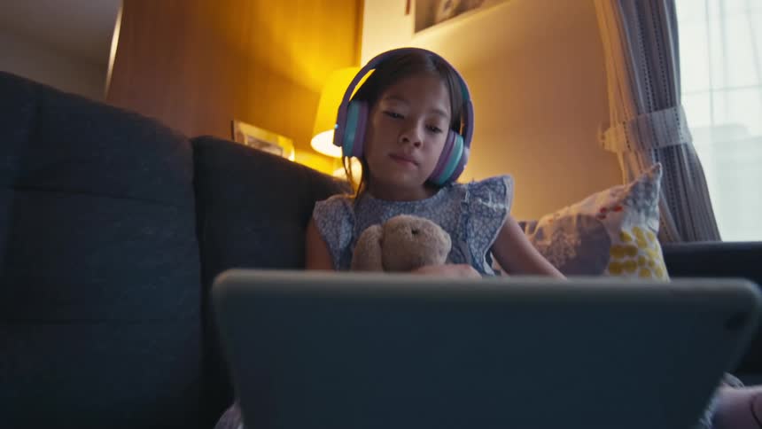 A young Asian girl wearing headphones sits on a cozy couch, holding her stuffed rabbit while watching a video on a tablet