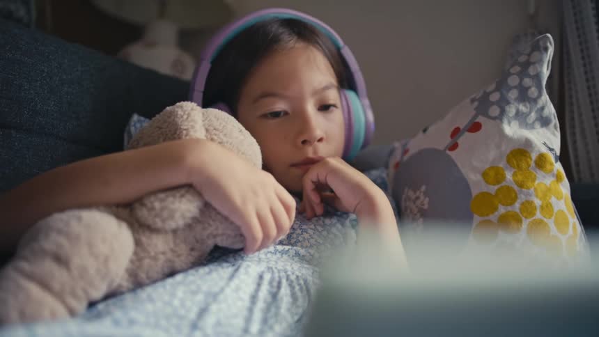 A young girl wearing headphones lies on the couch, cuddling her teddy bear while she watches a tablet