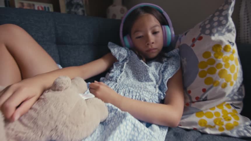 Young girl wearing headphones relaxes on a couch watching a tablet while holding a stuffed toy at home