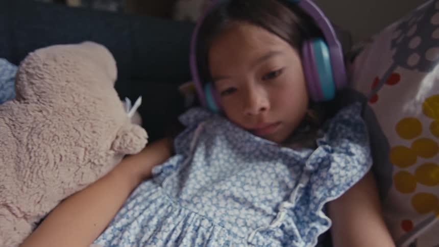 Close-up dolly shot of a serene young girl wearing headphones, captivated by her tablet while lounging on a sofa with her stuffed animal