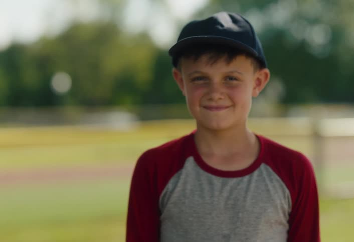 A happy young boy in a baseball cap smiles confidently for the camera while standing on a sunny baseball field