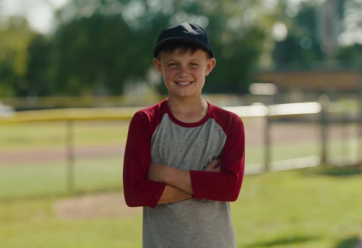 A happy young boy in a baseball cap and shirt smiles and crosses his arms while standing on a sunny baseball field