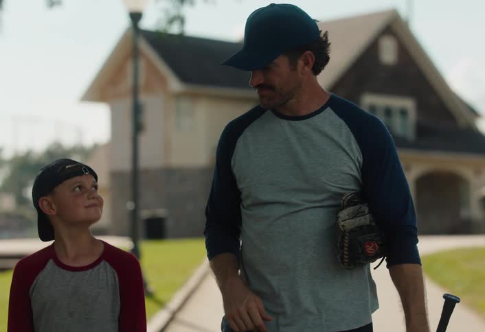 A father and his young son, dressed in baseball attire, walk and talk together on a path in a park on a sunny day