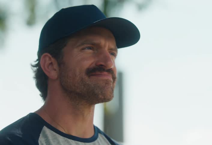 A content man with a mustache and a baseball cap smiles while enjoying a sunny day outdoors