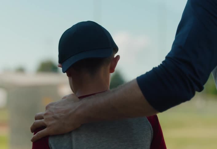 A father or coach offers a comforting pat on the head and shoulder to his young son as they walk together at a baseball field in slow motion