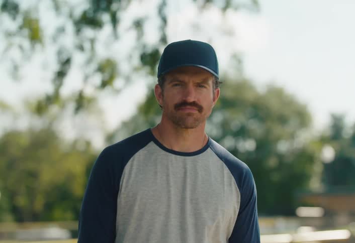 A serious baseball player with a mustache and cap looks up and stares intently into the camera on a sunny day