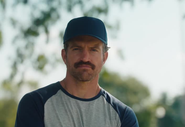 A serious man with a mustache wearing a baseball cap looks up with a determined expression while standing on a field