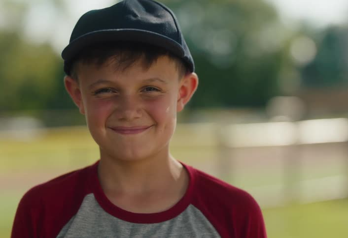 A young boy with dimples wearing a baseball cap smiles warmly at the camera in slow motion