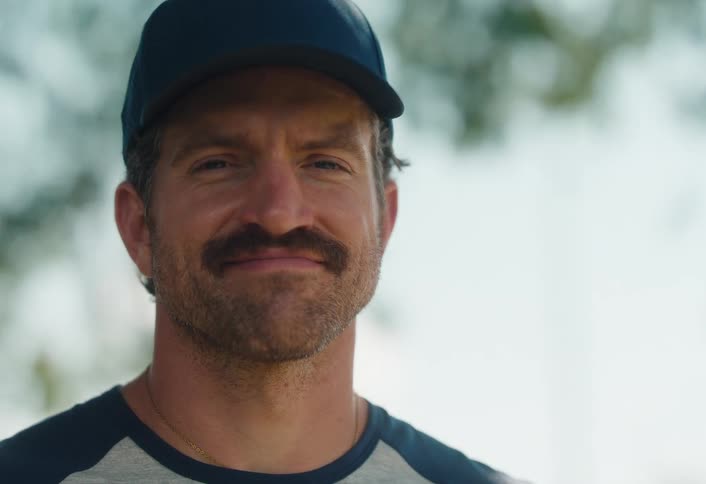 A confident man with a mustache and baseball cap looks to the side then turns his head to look directly into the camera with a calm smile