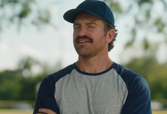 A proud and confident man with a mustache wearing a baseball cap smiles warmly for a portrait outdoors