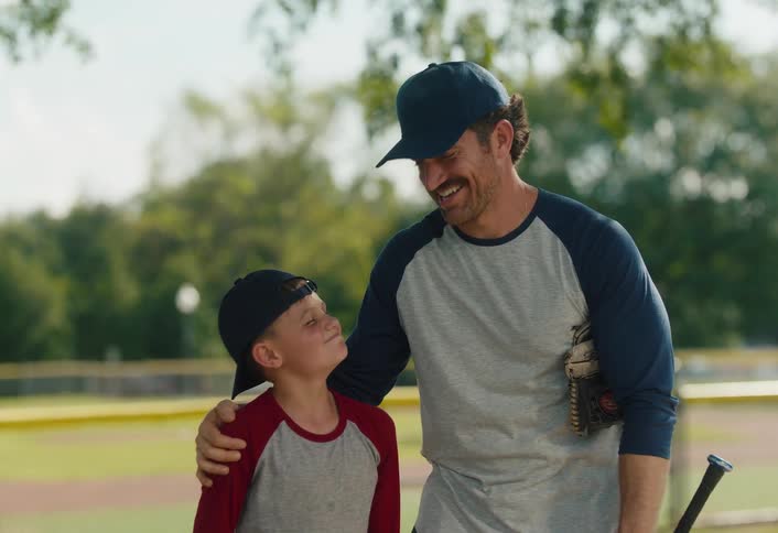 A smiling father and his son share a heartwarming moment while looking at each other on a sunny baseball field