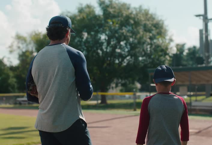 A father and son stand together at the edge of a baseball diamond, ready for a game of catch on a sunny afternoon