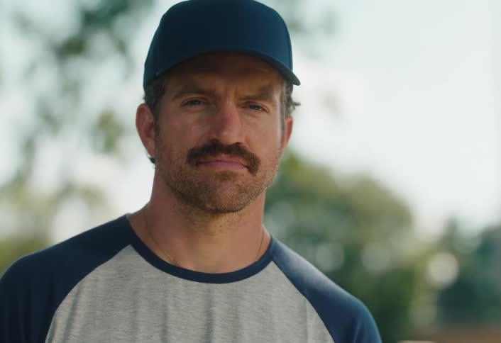 A confident man with a mustache and baseball hat looks directly into the camera with a serious expression that turns into a subtle smile