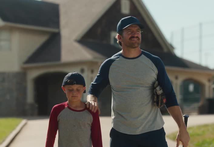 A proud father walks with his arm around his young son after baseball practice on a sunny day