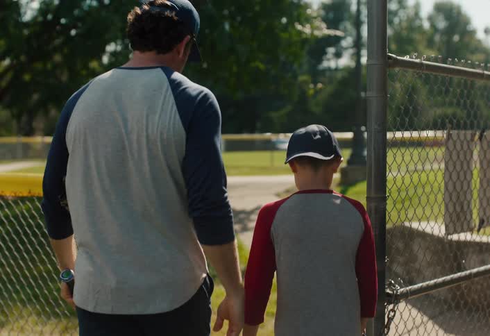 A supportive father puts his arm around his son as they walk off the baseball field together in slow motion