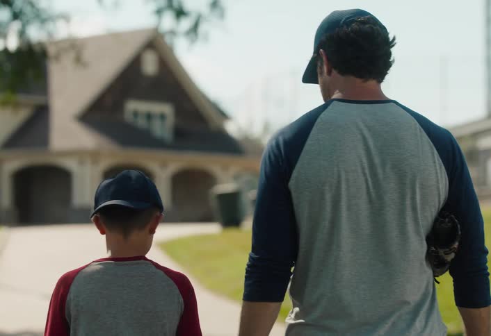 A father and son walk to a baseball field on a sunny day, with the father's supportive arm around his child