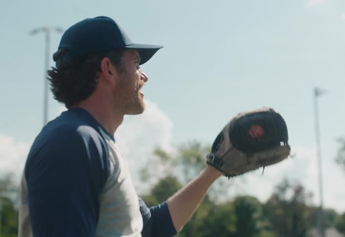 A man with a mustache smiles while throwing and catching a baseball in slow motion on a sunny day