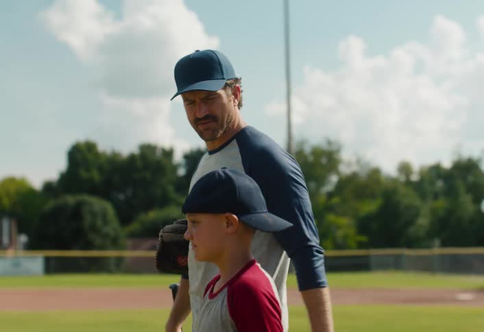 A father and his young son walk together across a baseball field on a sunny day