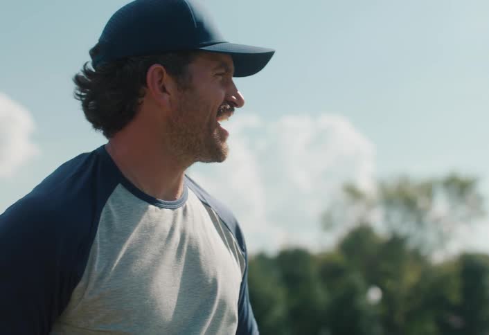 A smiling baseball player wearing a cap and glove throws a ball during practice on a sunny day in slow motion
