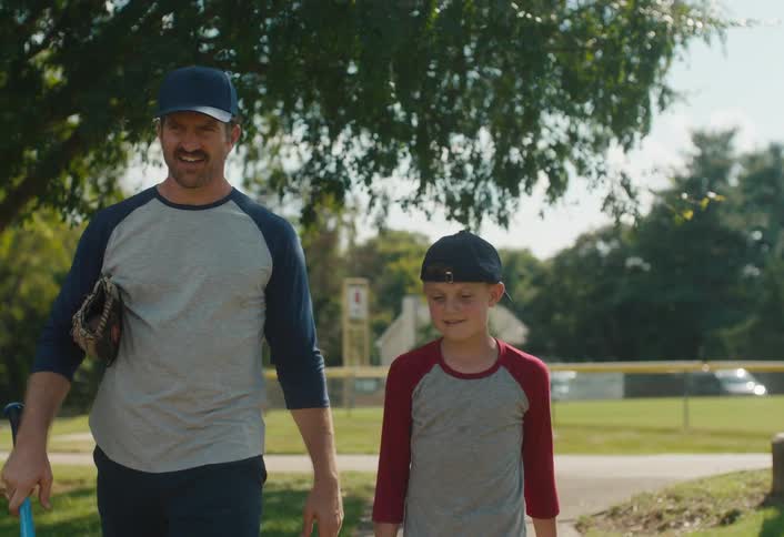 A father and son share a bonding moment as they walk through a park with their baseball equipment in slow motion