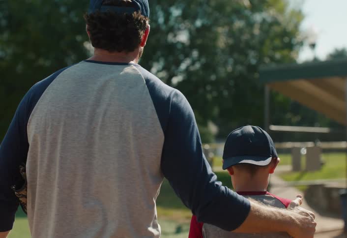 A supportive father puts his arm around his son as they walk together on a sunny baseball field