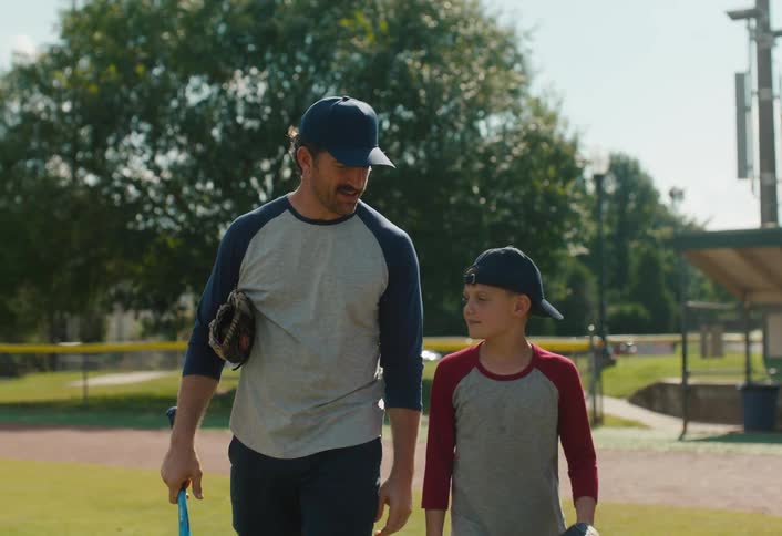 A father and son walk and talk together on a baseball field, sharing a fist bump in slow motion on a sunny day