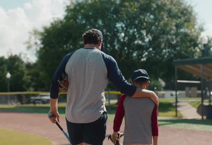 A father puts his arm around his son as they walk together with their baseball gear at a park in slow motion