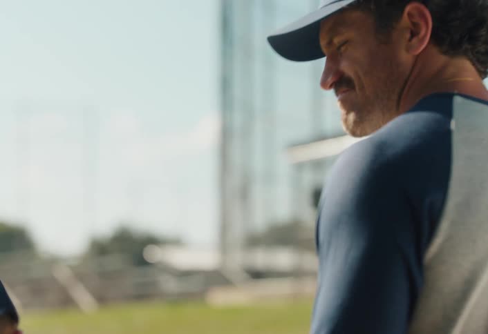 A father places a supportive hand on his young son's shoulder as they walk together towards a baseball field in slow motion