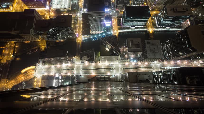 A dizzying top-down timelapse view of a sprawling city at night, with traffic light trails weaving between illuminated skyscrapers