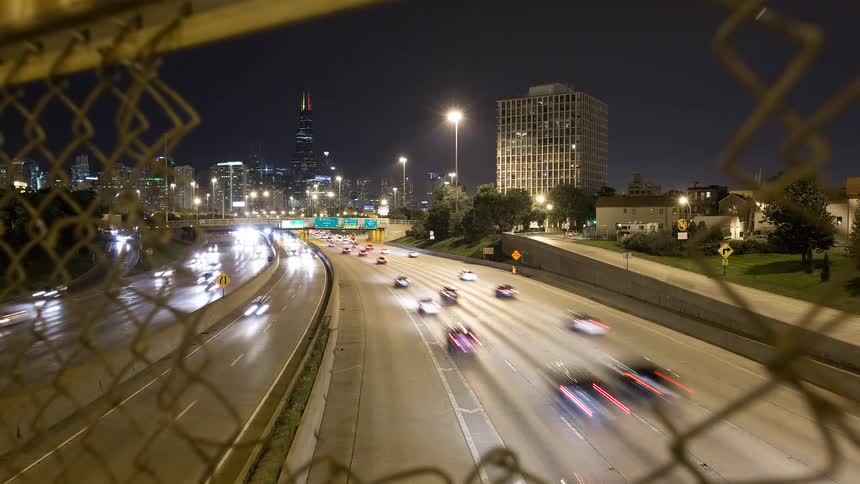 A dynamic dolly-out timelapse captures streaks of traffic light on a bustling Chicago highway at night, viewed from an overpass through a chain-link fence