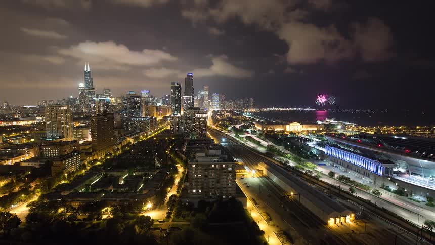 An elevated time-lapse view of the Chicago skyline at night shows bustling traffic and train activity while celebratory fireworks explode over Lake Michigan