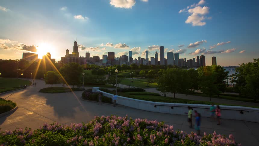 A vibrant sunset timelapse over the Chicago skyline as people enjoy an evening stroll through a beautiful park with blooming flowers in the foreground