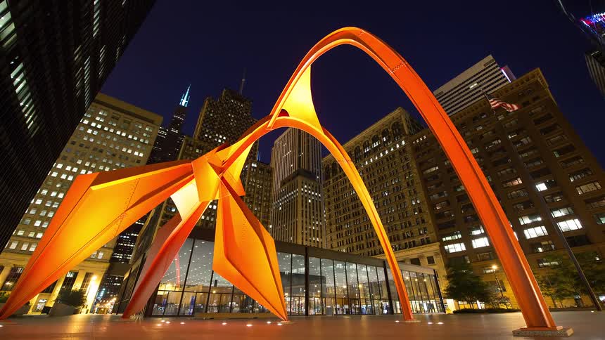 A slow zoom-out timelapse of a vibrant orange sculpture set against the illuminated Chicago skyline at night