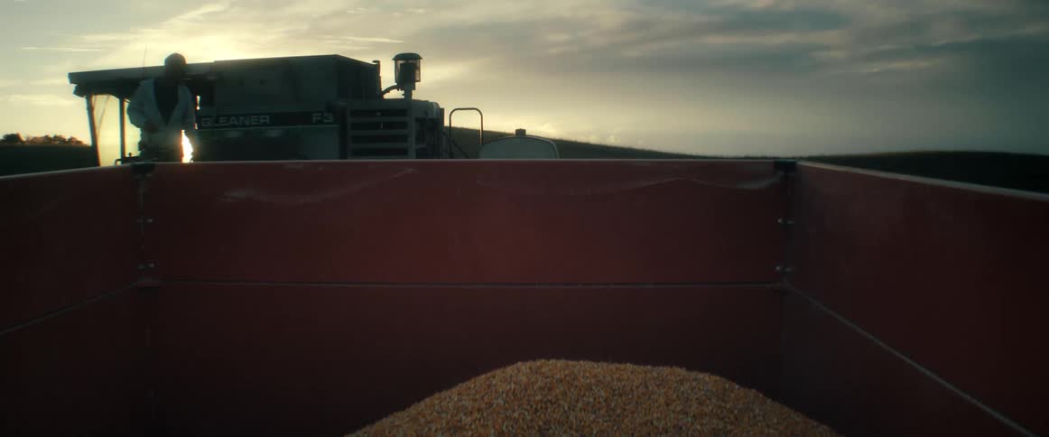 A combine harvester is silhouetted against the setting sun as seen from inside a wagon full of freshly harvested corn
