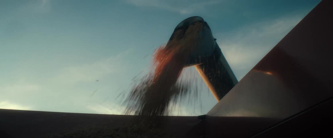 A stream of golden grain flows from a combine harvester auger into a hopper during a beautiful sunset harvest