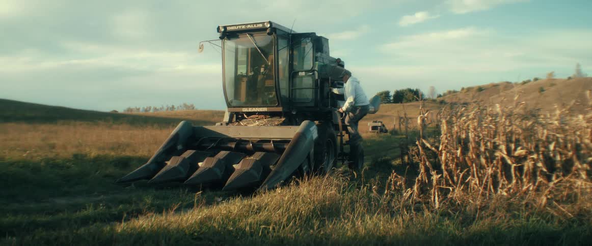A male farmer climbs up into the driver's cab of a large combine harvester parked in a rural field