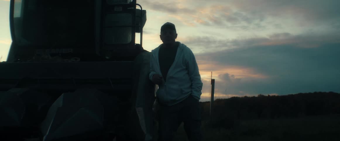 A farmer stands silhouetted against the sunset, leaning on his combine harvester after a long day in the field