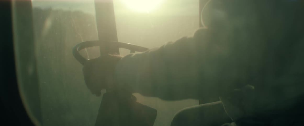 A farmer's hands grip the steering wheel inside a dusty tractor cab during a hazy sunrise