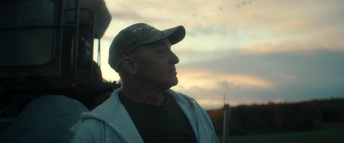 A contemplative senior farmer wearing a cap looks out over his field during a beautiful sunset
