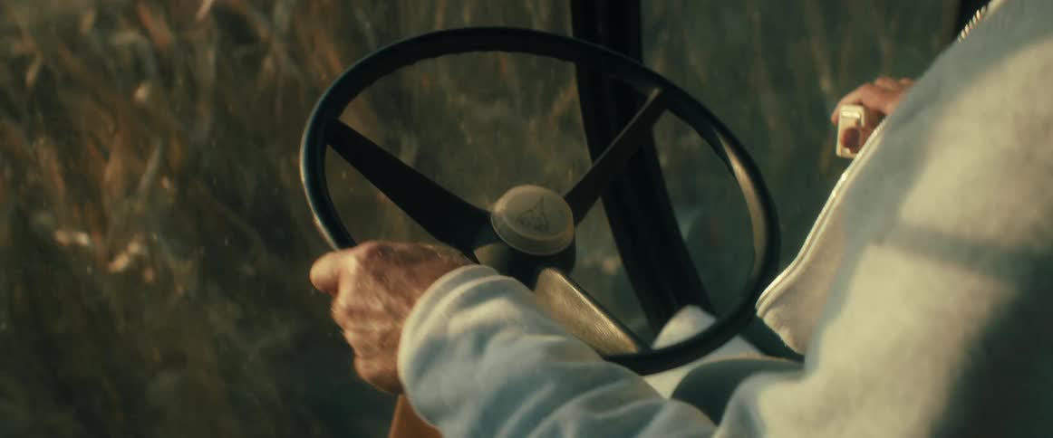 A farmer's hands grip the steering wheel of a farm vehicle, navigating through a golden cornfield at sunset during harvest