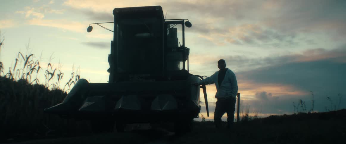 A silhouetted farmer stands proudly next to his combine harvester in a field at dusk