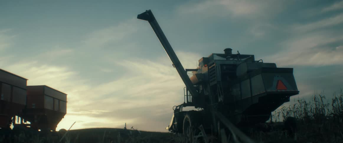 A cinematic low-angle shot shows a combine harvester extending its auger to unload grain at sunset in a rural cornfield
