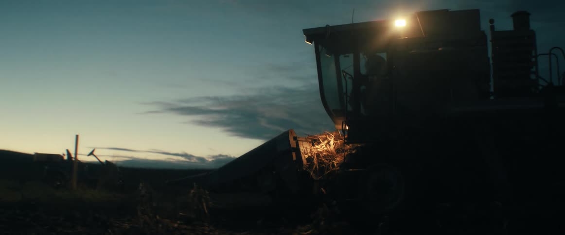 A silhouetted farmer operates a combine harvester, its lights cutting through the twilight as it moves through a vast agricultural field