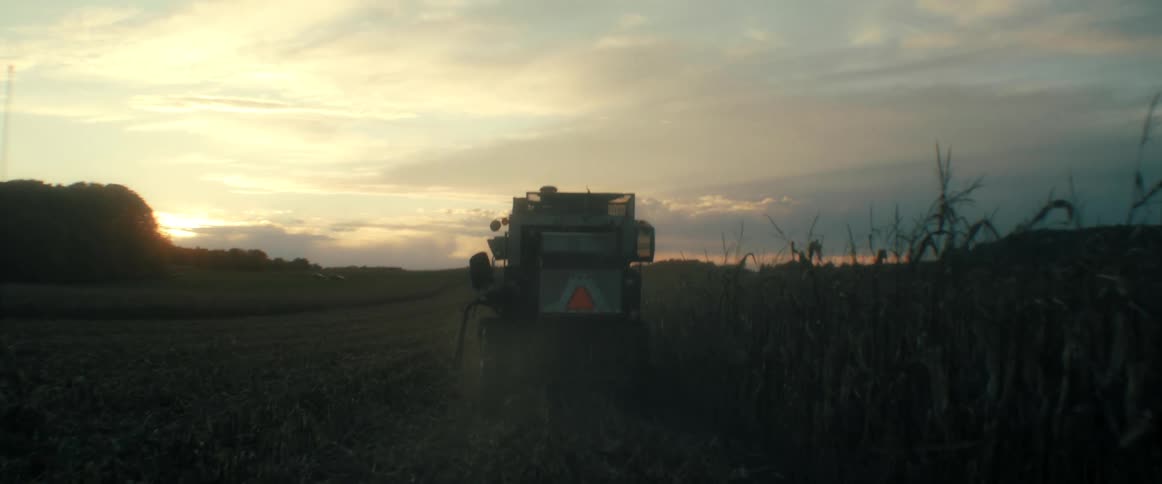 A combine harvester drives through a rural cornfield at sunset, kicking up dust as it works