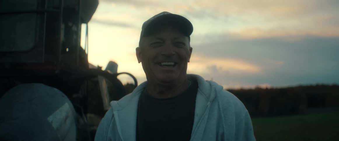 A happy, smiling older farmer wearing a camouflage cap laughs while standing in a field next to his tractor at sunset