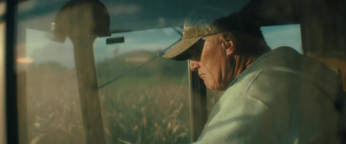 A thoughtful older farmer operates a combine harvester, driving through a sunlit field of crops