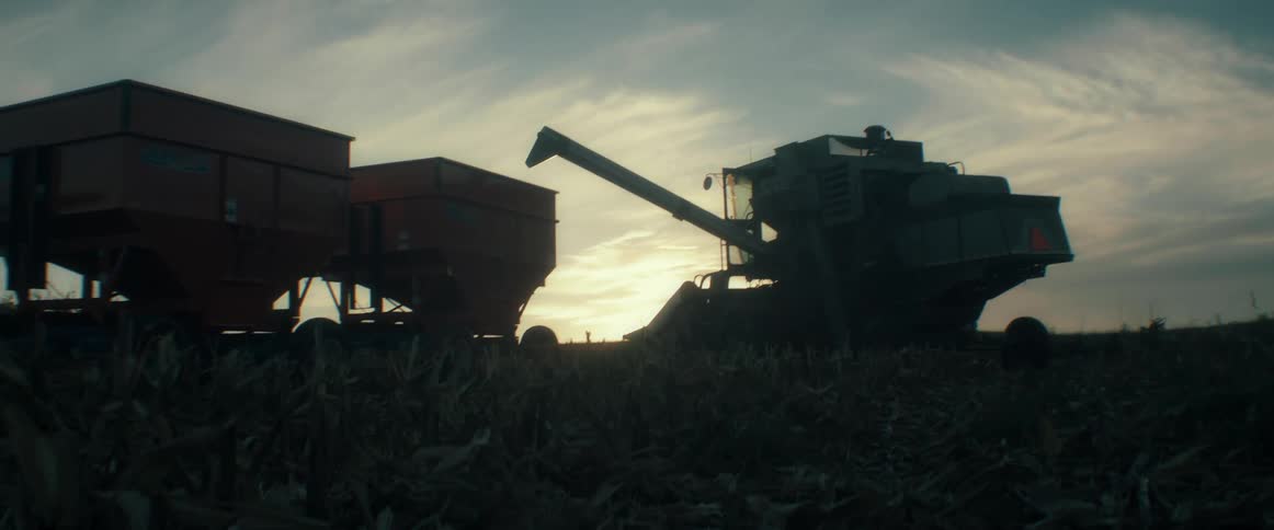 A combine harvester transfers freshly harvested corn into a grain cart as the sun sets over the field